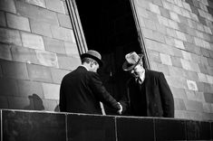 two men in suits and hats are shaking hands on the side of a brick building