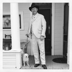 a man in a suit and hat is standing on the porch with his small dog