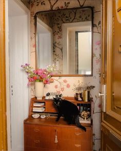 a black cat sitting on top of a dresser next to a mirror and vase with flowers