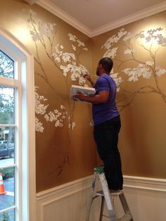 a man is painting a wall with flowers on it and another man is standing on a ladder in front of the window