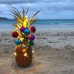 a pineapple decorated with christmas balls and lights on the sand at the beach in front of an ocean