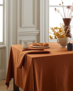 an orange table cloth with croissants and flowers on it in front of a window