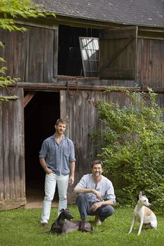 two men are standing in front of a barn with three dogs and one dog is sitting on the grass