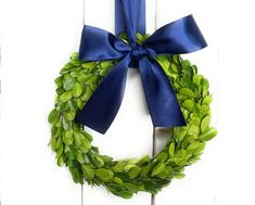 a green wreath with blue ribbon hanging on a white wooden wall, decorated with greenery