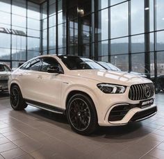 a white mercedes suv is parked in front of large windows and glass walls with other cars behind it