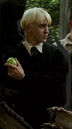 two young men sitting next to each other in front of trees and one holding an apple