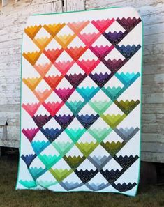 a colorful quilt hanging on the side of a white wall next to a fence and grass