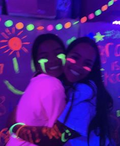 two young women are smiling and hugging in front of neon lights on the wall behind them