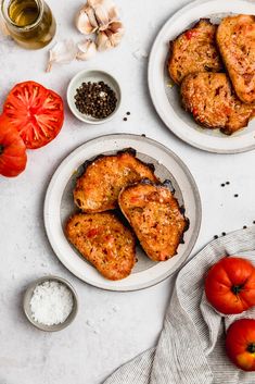 two white plates topped with meat next to tomatoes and garlic