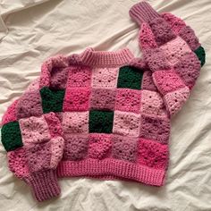 a pink and green knitted sweater laying on top of a white bed covered in sheets