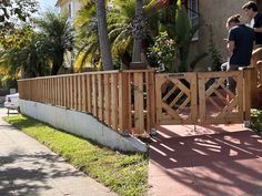 two people standing on the side of a building near a wooden fence and palm trees