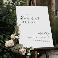 a sign with flowers on it sitting in front of some bushes and trees at night before an outdoor wedding