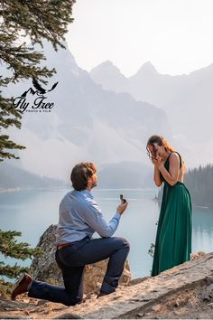 a man kneeling down next to a woman on top of a cliff near a lake