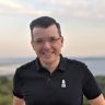 a man in black shirt and glasses standing on top of a hill next to the ocean