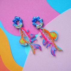 Laser cut fish earrings featuring a unique hand painted pattern of orange, pink, yellow, purple, blues and gold glitter resin with tiny hand painted details. The fish dangle from a small blue and gold glitter top piece.- Painted pattern is over clear, see through, acrylic. This effect gives the earrings a fun layered depth.- Earrings are 2.75 inches long by 1.25 inches wide. - Earrings are push back studs in hypoallergenic stainless steel. - Lightweight, not heavy at all. Great for daily wear.- Hand Painted Pattern, Mixed Media Jewelry, Glitter Top, Orange And Gold, Plastic Crafts, Tiny Hand