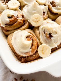 a white dish filled with cinnamon rolls covered in icing and sliced banana's