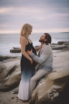 a man and woman are sitting on rocks near the ocean, looking at each other