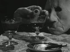 a creepy man holding an eyeball in front of two empty glasses on a table