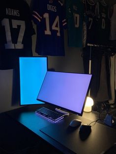 a computer desk with two monitors and a keyboard on it in front of some jerseys