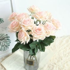 a vase filled with pink roses sitting on top of a white rug next to an open book