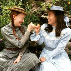 two women in dresses and hats are sitting on a bench eating fruit together, with one woman smiling at the camera