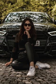 a woman sitting on the ground in front of a black car with her hand under her chin