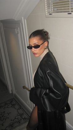 a woman in black leather jacket and sunglasses standing next to a wall with an open door