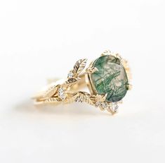 This exquisite Moss Agate ring set features the serene beauty of nature captured in elegant simplicity. Crafted with care, it showcases the captivating charm of moss agate, a stone renowned for its lush green hues and intricate patterns reminiscent of verdant forests. The delicate leaf motif adorning the band adds a touch of organic grace, enhancing the ring's natural charm. Elegant Moss Agate Rings With Natural Inclusions, Green Moss Agate Jewelry With Natural Stones, Nature-inspired Yellow Gold Jewelry With Gemstone, Green Emerald Jewelry With Rose Cut Diamonds, Nature-inspired May Birthstone Jewelry, Oval Green Jewelry With Rose Cut Diamonds, Green Rose Cut Diamond Rings As A Gift, Moss Agate Oval Wedding Rings, Elegant Oval Moss Agate Rings