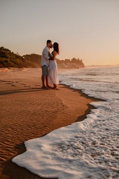 a man and woman standing on top of a beach next to the ocean at sunset