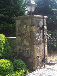 an iron gate with a light on top and stone wall around it in front of some bushes