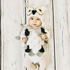 a baby dressed up as an owl standing in front of a white wall with wood planks