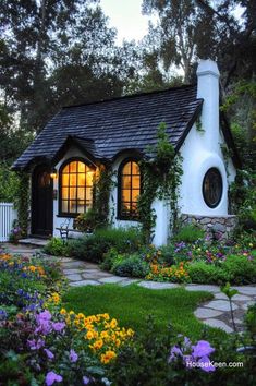 a small white house surrounded by flowers and greenery in the evening light, with a stone pathway leading to it