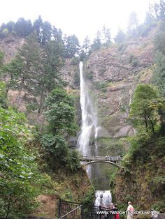 people are standing at the bottom of a waterfall with a bridge in front of it