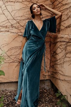 a woman standing in front of a wall wearing a blue dress and holding her hand on her head