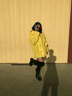 a woman in yellow coat and black boots standing next to a wall with her shadow on the ground