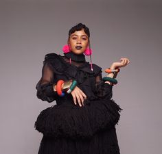 a woman in a black dress with pink earrings and beads on her hands, holding two colorful objects