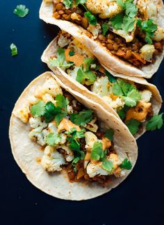 three tacos filled with meat and vegetables on top of a black surface, topped with cilantro