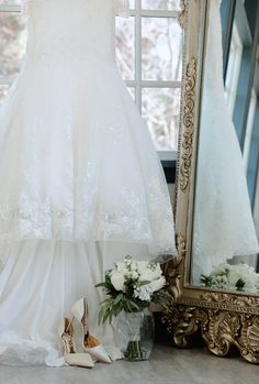 a wedding dress hanging in front of a mirror next to flowers and shoes on the floor