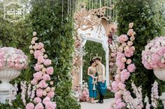 two people standing in front of a garden with pink and white flowers on the walls