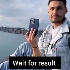 a man holding up his cell phone in front of the water and bridge that reads wait for result