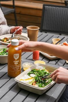 two people sitting at an outdoor table eating salad and condiments on the side