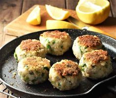 four crab cakes in a pan with lemons on the side and a cutting board behind them