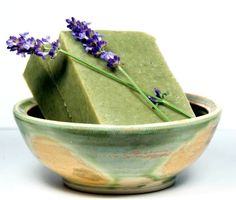 a green soap bar in a bowl with lavender flowers on the top, and a single purple flower sticking out of it