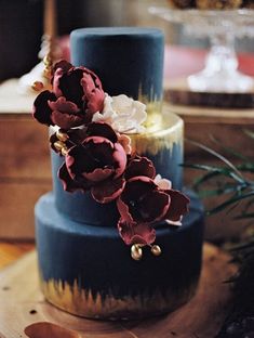 a three tiered cake with flowers on the top is blue and has gold trim