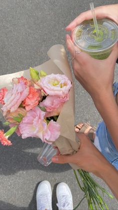 a woman holding flowers and a drink in her hand while sitting on the ground next to another person