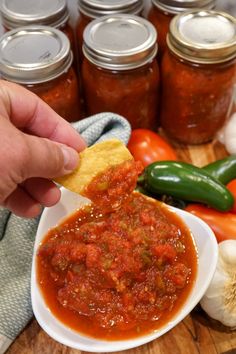 a hand holding a tortilla chip over a bowl of salsa with peppers and garlic on the side