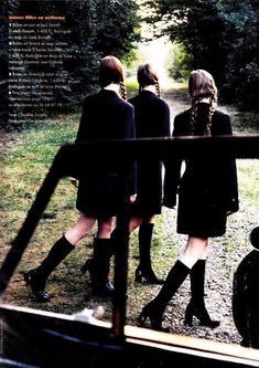 three girls in black dresses and boots are looking at something through a window with trees behind them