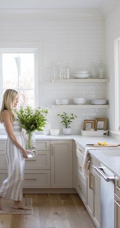 a woman standing in a white kitchen holding a potted plant and looking out the window