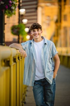 a young man leaning against a yellow fence smiling at the camera with his hands on his hips