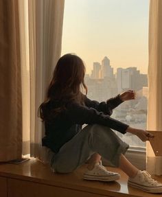 a woman sitting on top of a window sill looking out at the city skyline